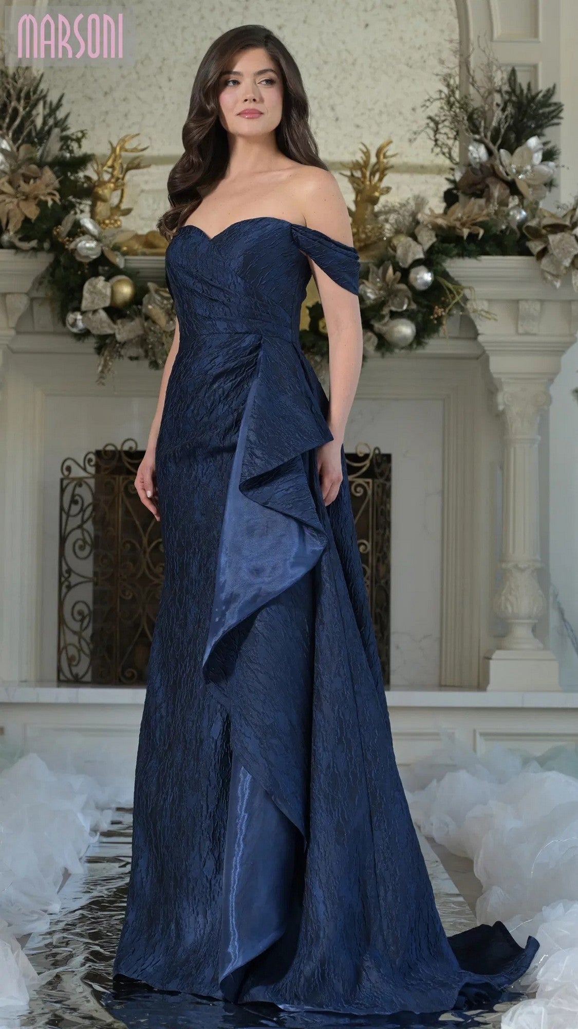 A woman stands indoors in the Marsoni MV1365 long formal dress, a navy off-the-shoulder sheath gown with a ruched bodice and side draping. Ornate decor and floral arrangements adorn the background.